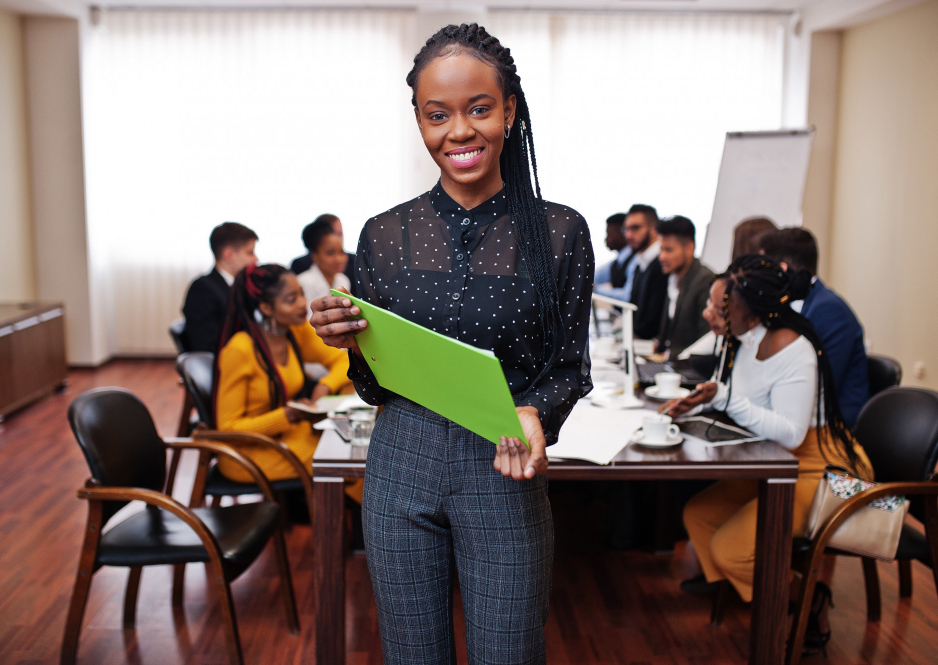 Black business woman smiling with braids holding green clipboard in front of a table full of people having a meeting
