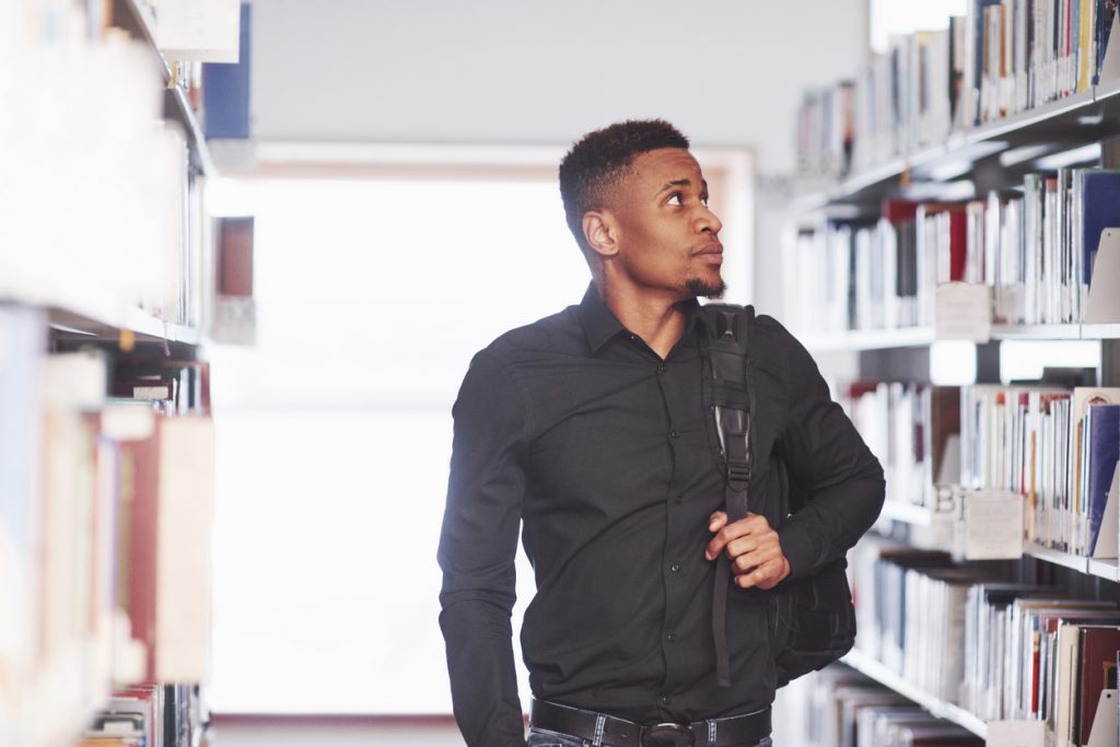 Walking forward with backpack. African american man in the library searching for some books.