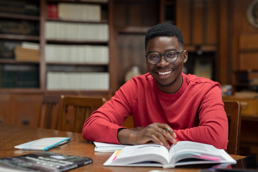 Portrait of university student doing homework in school library and smiling. Happy high school student looking at camera while studying for exam. African american clever guy with open book sitting at desk with copy space.