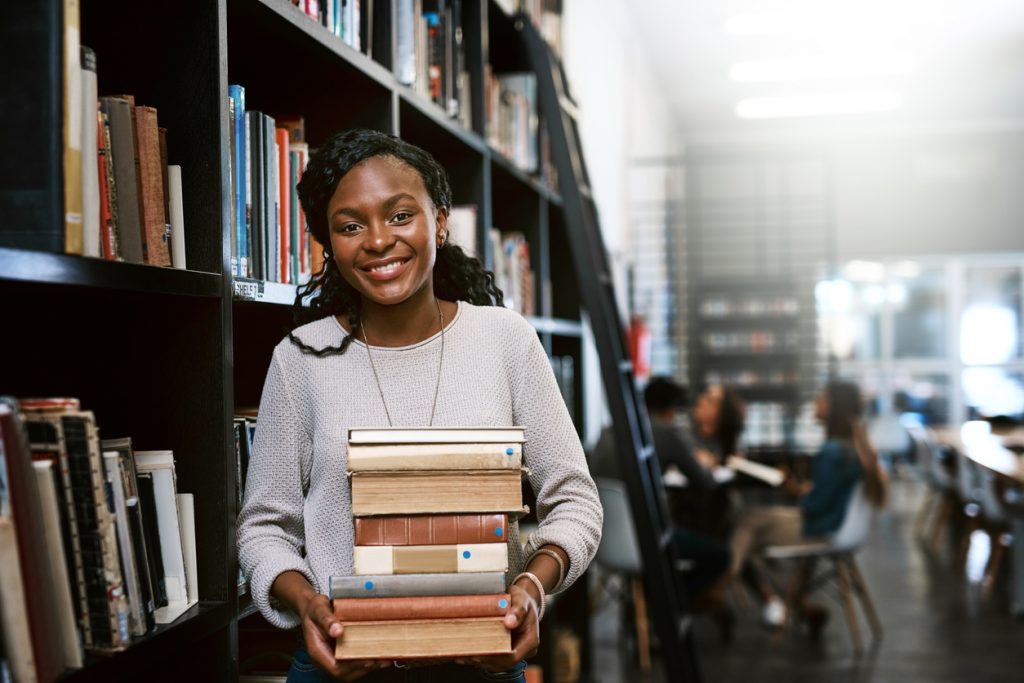 Portrait of a happy young black woman carrying books in a library at college